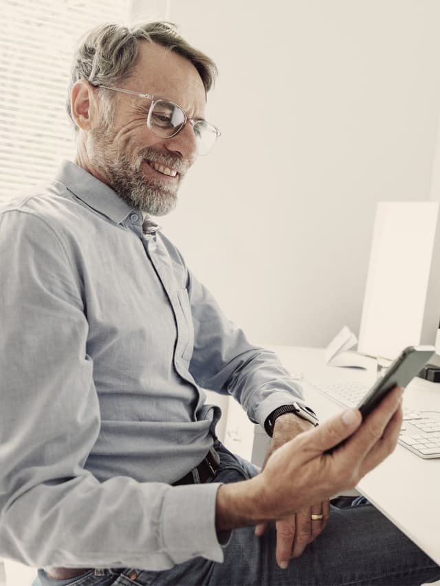 Eine Person sitzt an einem Schreibtisch, hält ein Smartphone in der Hand und schaut darauf. Die Person trägt ein hellblaues Hemd und eine Brille, vor der Person steht ein Computermonitor.