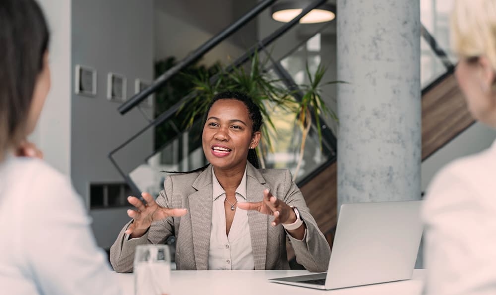 Eine Frau in einem beigen Anzug gestikuliert ausdrucksstark während einer Besprechung. Sie sitzt an einem Tisch mit einem Laptop und unterhält sich mit zwei Personen, was eine Atmosphäre der Zusammenarbeit vermittelt.