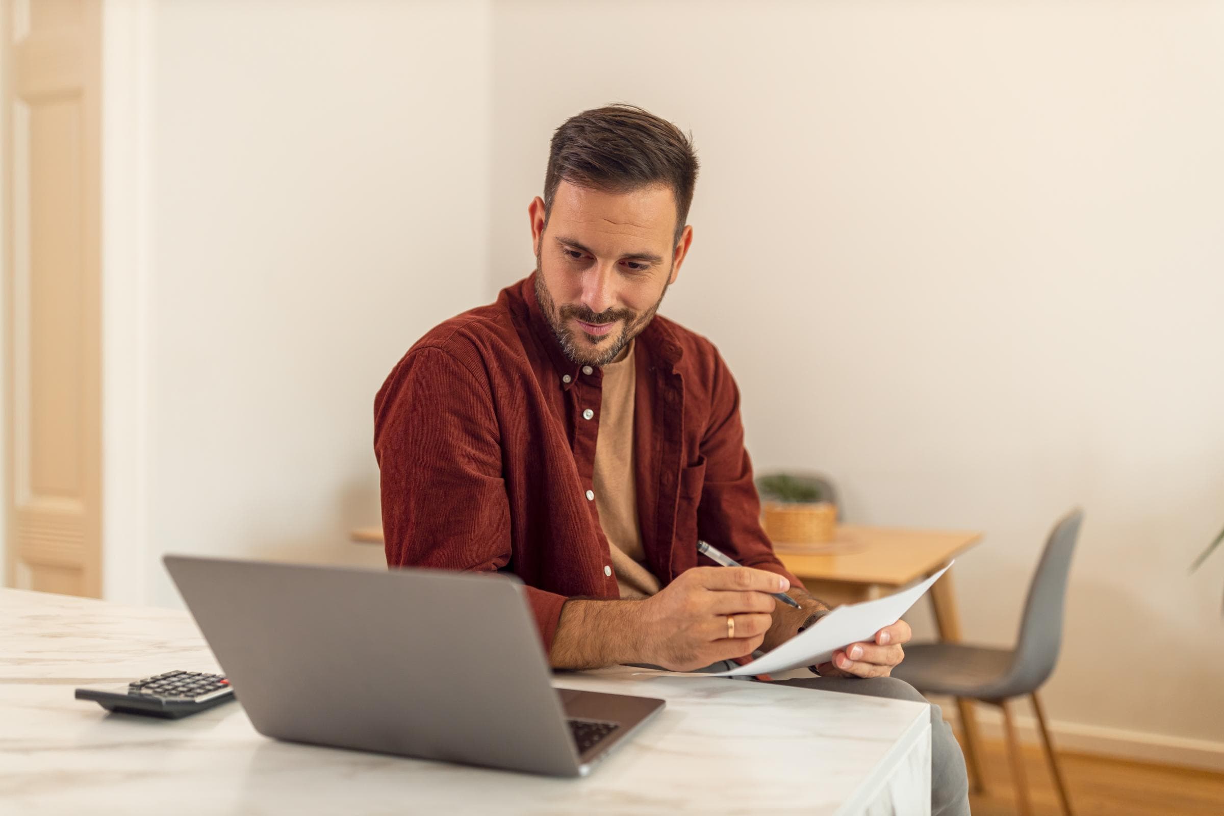 Ein Mann im mittleren Alter mit braunen Haaren und dunkelrotem Hemd sitzt vor seinem Laptop am Tisch und macht sich Notizen. Neben dem Laptop liegt ein Taschenrechner.