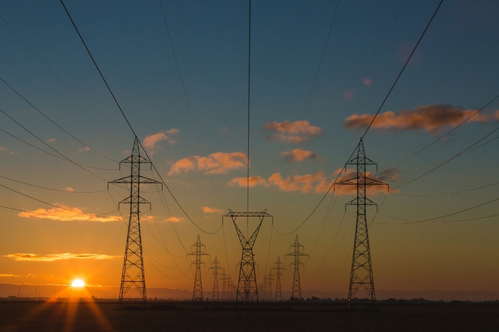 Fotografie von Hochspannungsmasten bei Sonnenuntergang mit orangefarbenem Himmel und vereinzelten Wolken. Stromleitungen verlaufen symmetrisch von vorderen zu hinteren Masten, was Tiefe und Perspektive betont.