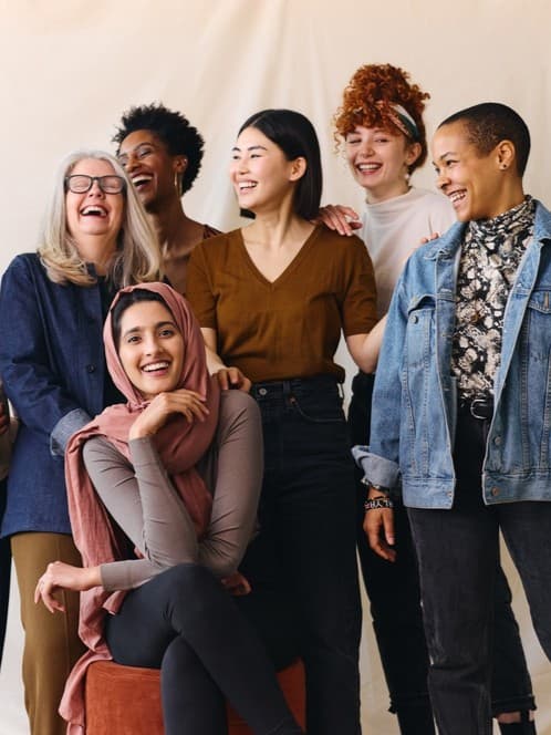 Sieben Frauen unterschiedlicher Herkunft stehen zusammen, lächeln und lachen. Sie strahlen in einer ungezwungenen, freundlichen Umgebung ein Gefühl der Zusammengehörigkeit und Freude aus. Sie repräsentieren gründende Frauen.