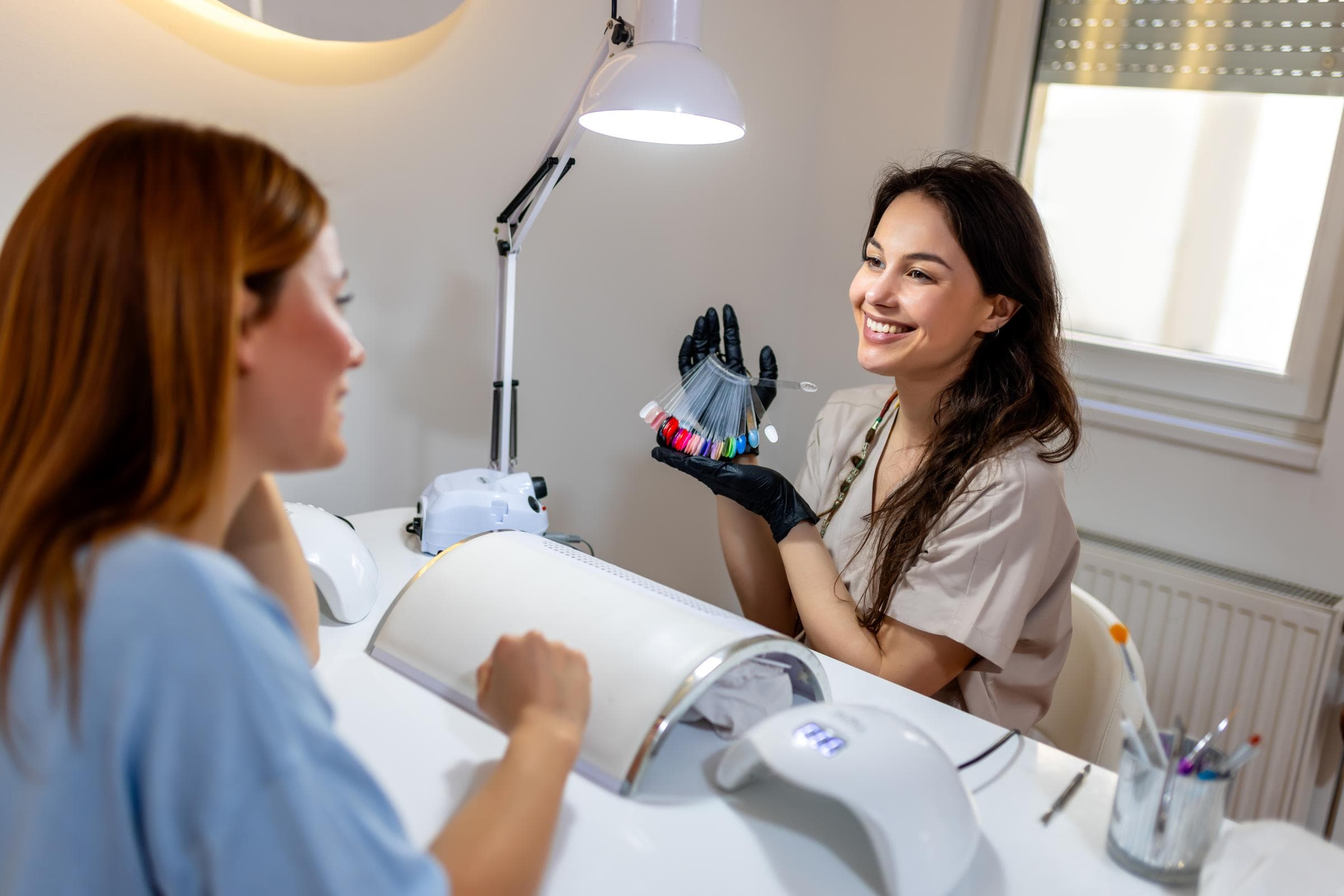 Eine lächelnde Nageltechnikerin zeigt einer Kundin an einem gut beleuchteten Manikürtisch Farbmuster. Die Umgebung ist hell und einladend und vermittelt eine fröhliche Atmosphäre.