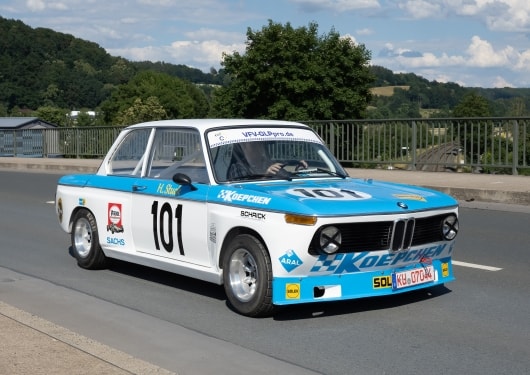 Ein Oldtimer-Rennwagen BMW 2002 mit blau-weißer Lackierung und der Nummer 101 fährt auf einer Straße. Er ist umgeben von grünen Hügeln unter einem teilweise bewölkten Himmel.