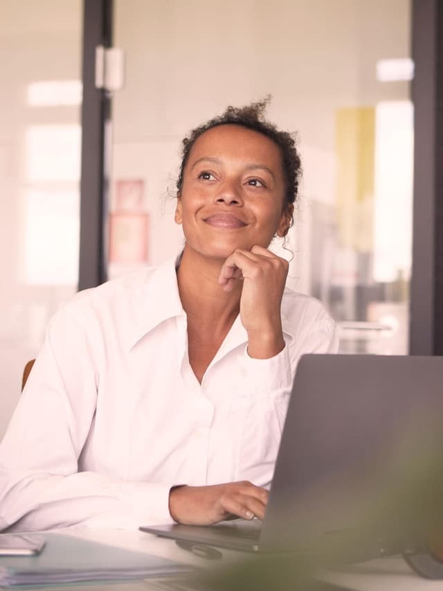 Der Blick einer lächelnden Frau in einem weißem Hemd ist nach oben gerichtet und auf dem Schreibtisch vor ist ein Laptop.