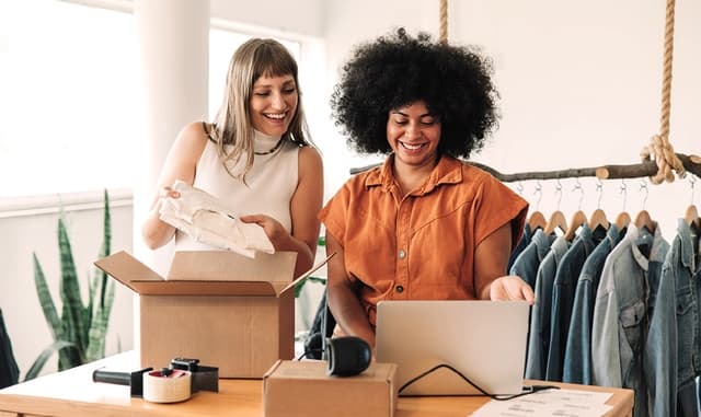 Zwei junge Frauen packen Kartons für den Versand. Sie stehen in ihrem privaten Lager mit einem Kleiderständer und einer Packstation. Die Frau recht trägt eine orange Bluse, hat stark gelocktes schwarzes Haar und lächelt. Die Frau links hat ein weißes Oberteil an und blonde Haare.