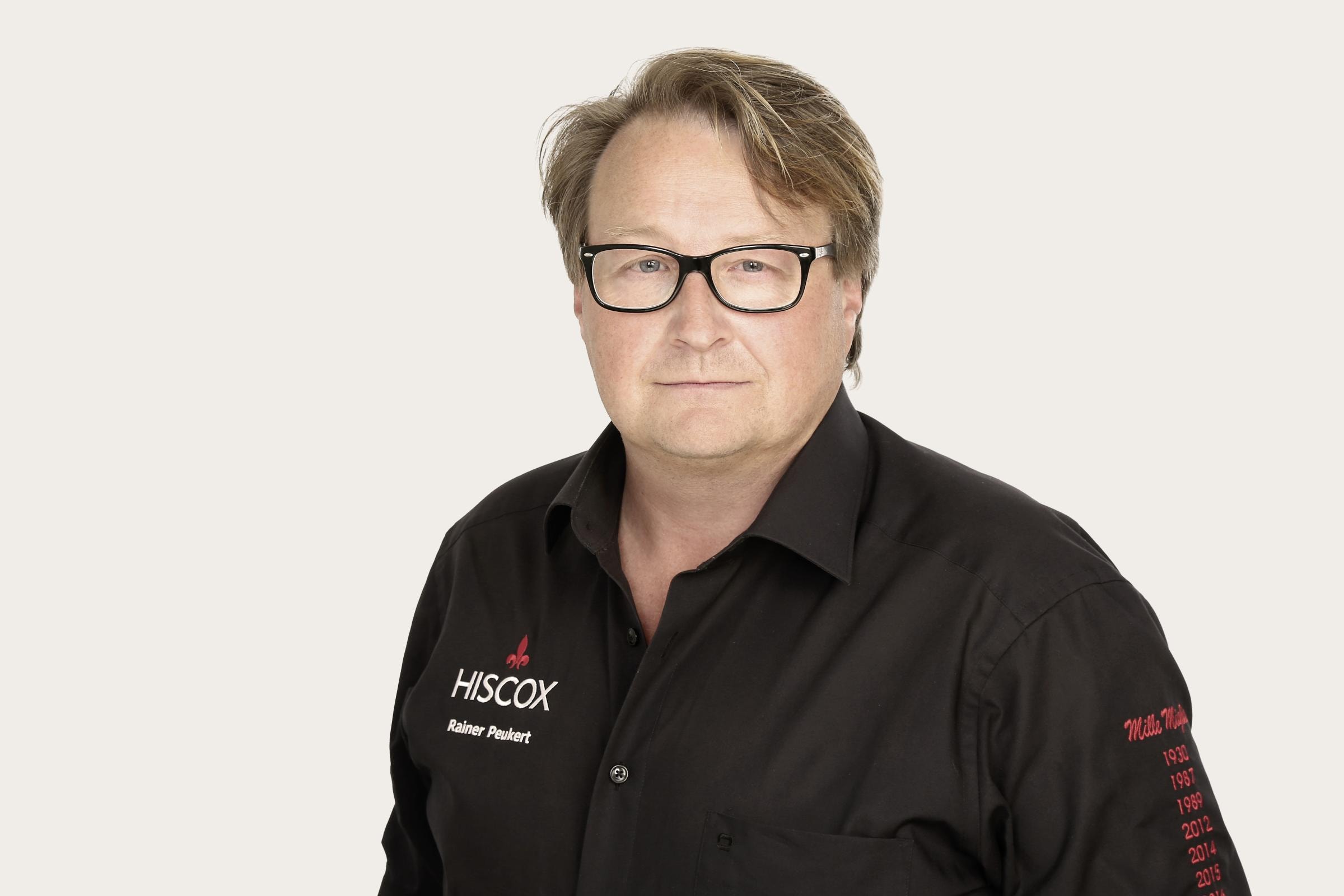 Rainer Peukert ein Oldtimer-Experte bei Hiscox trägt einem schwarzem HISCOX-Hemd, Namensstickerei ‚Rainer Paukert‘ auf der Brust und roten Jahreszahlen auf dem rechten Ärmel.