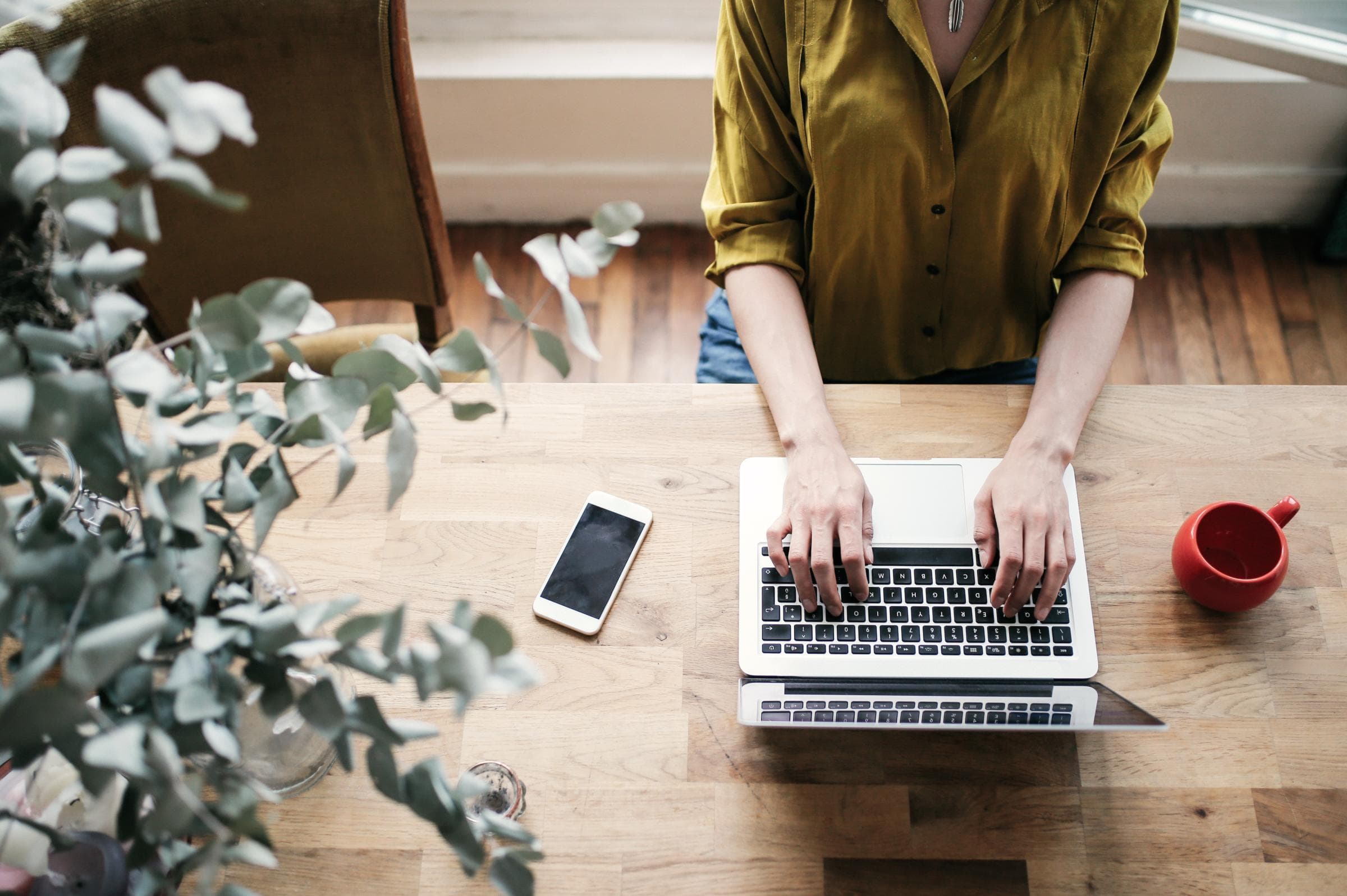 Person tippt auf einem Laptop an einem Holztisch mit Smartphone, roter Tasse und Pflanze.