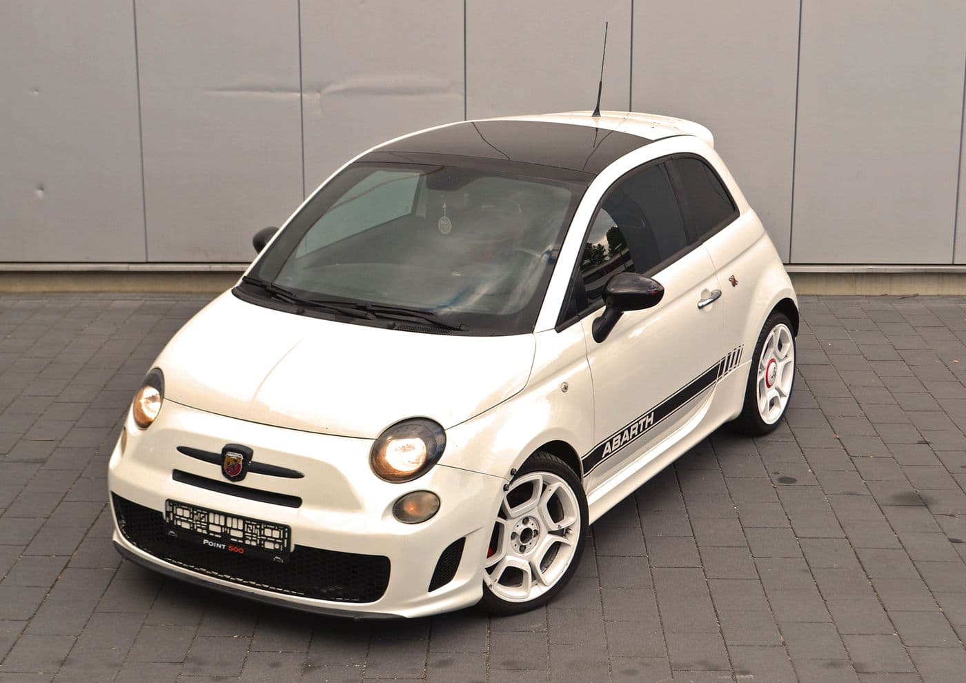 Schöner weißer Abarth 500 mit schwarzen Details in der Frontansicht parkend vor einer Wand mit angeschalteten Scheinwerfern.