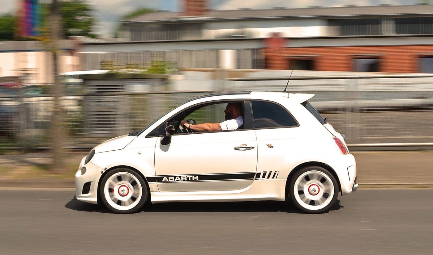 Weißer Abarth 500 mit schwarzen Details ist bei Sonnenschein fahrend auf einer Straße. Man sieht ihn von der Seitenansicht mit männlichem Fahrer hinterm Steuer.