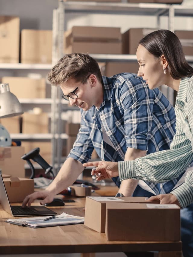 Ein Mann mit Brille und Hemd und eine junge Frau mit brauen Haaren stehen an einem Packtisch und bearbeiten Bestellungen an einem Laptop.