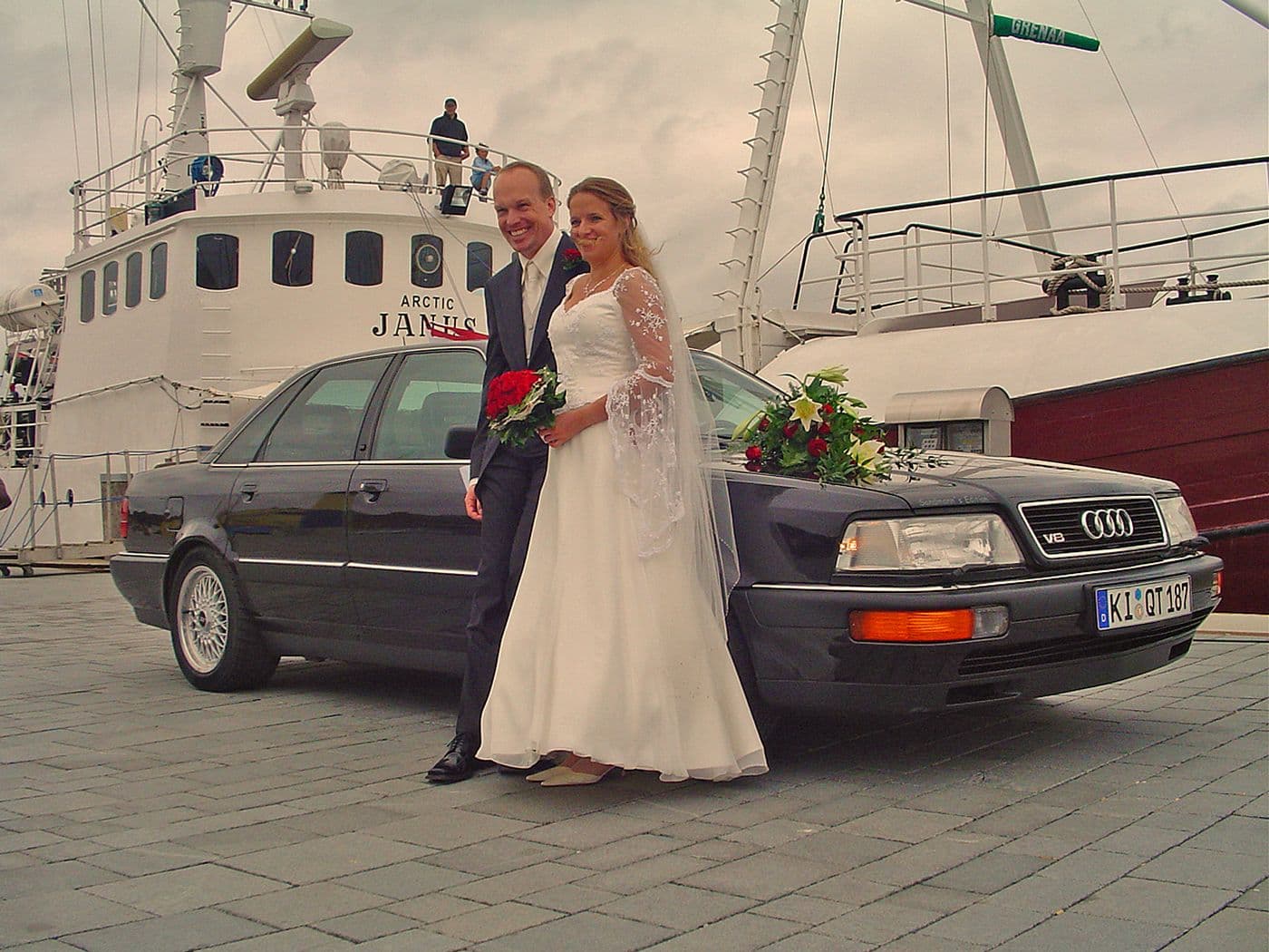 Brautpaar steht vor dem Audi v8 4,2 Quattro, der an einem Hafen vor einem Schiff parkt. Das Brautpaar lächelt und ist an das Auto gelehnt.