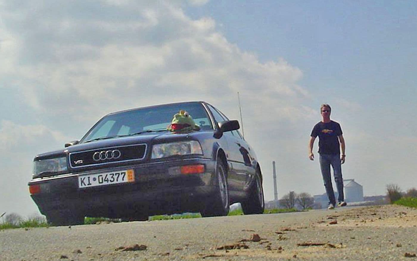 Jens Tanz geht auf einer Straße auf seinen schwarzen Audi v8 zu. Man sieht den Oldtimer von der Frontansicht, auf der Motorhaube liegt ein Kermit-Kuscheltier.