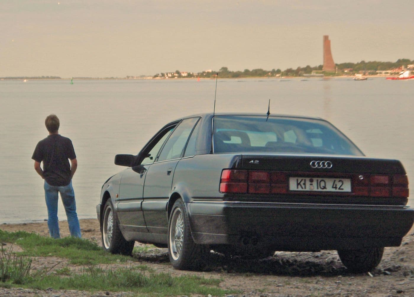 Jens Tanz steht mit dem Rücken zur Kamera auf Ufer eines Sees nehmen einem schwarzen Audi v8.