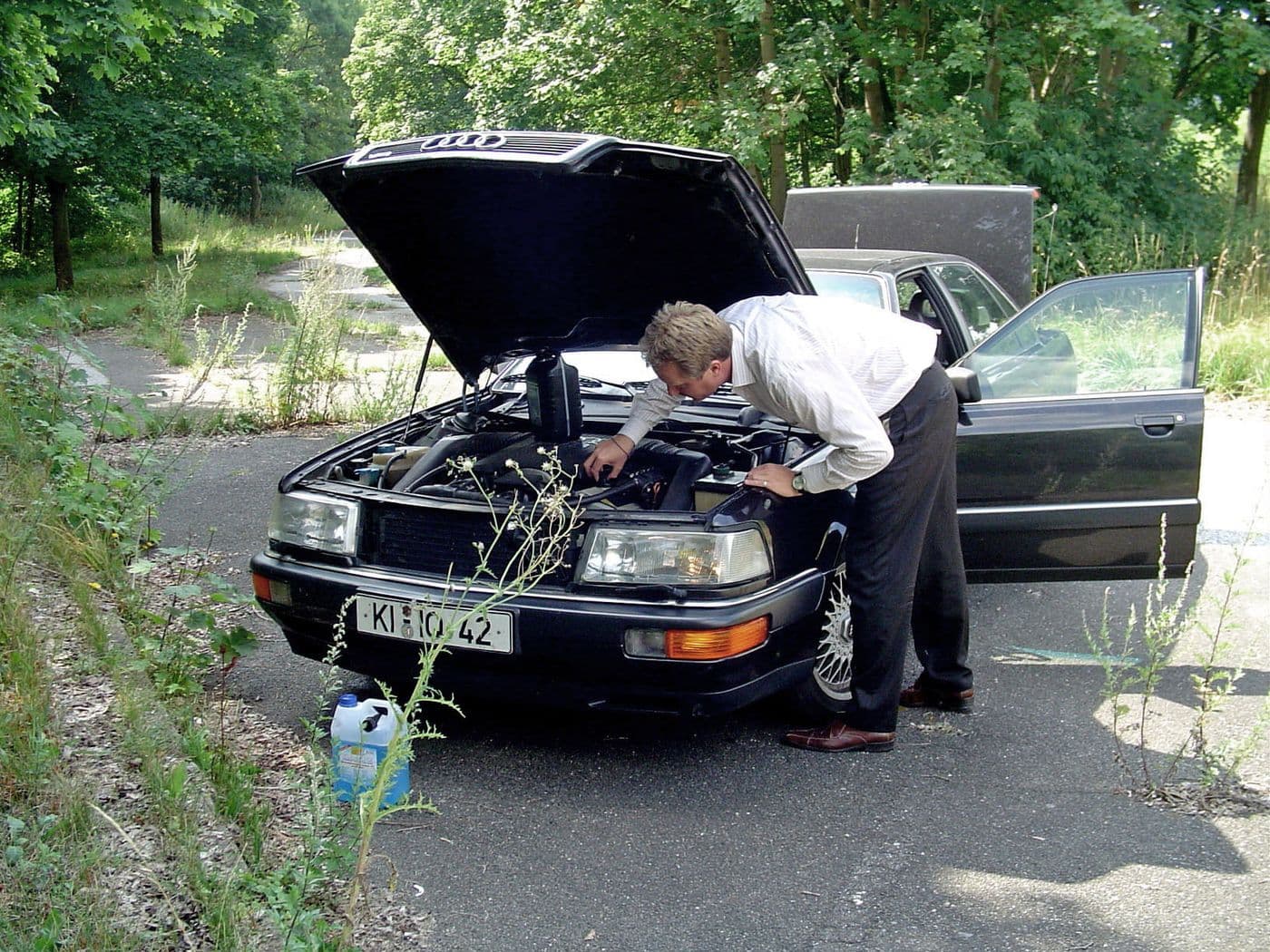 Jens Tanz blickt unter die Motorhaube eines schwarzen Audi v8. Das Auto parkt auf dem Teer, umgeben von grünen Bäumen, Motorhaube, Türen und Kofferraum sind geöffnet.