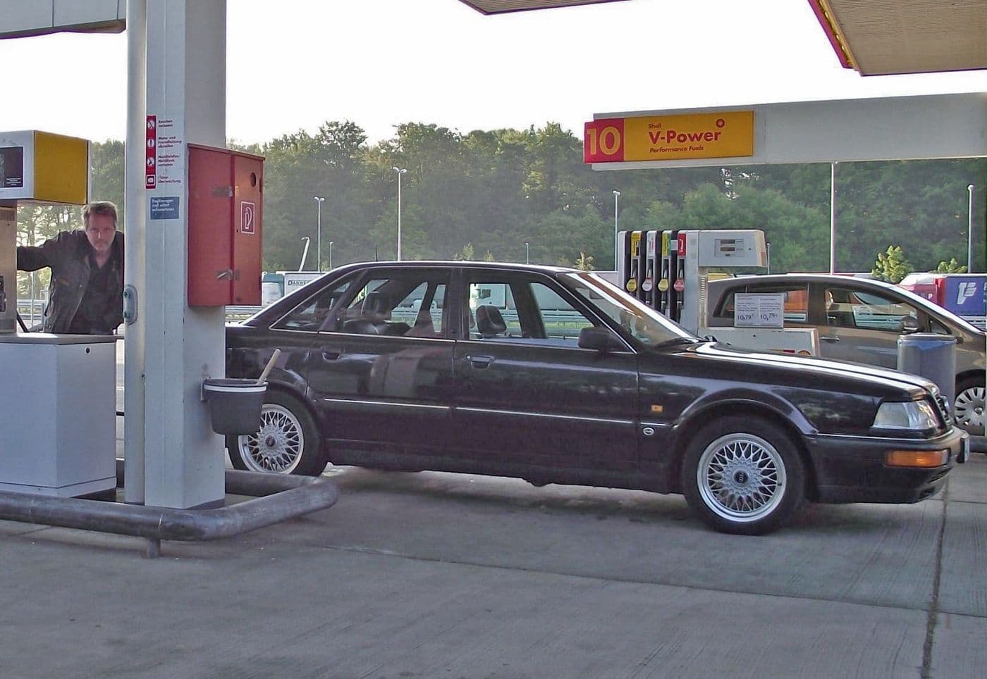 Der schwarze Audi v8 steht an der Tankstelle und wird getankt. Man sieht das Fahrzeug parkend von der Seite neben der Zapfsäule.