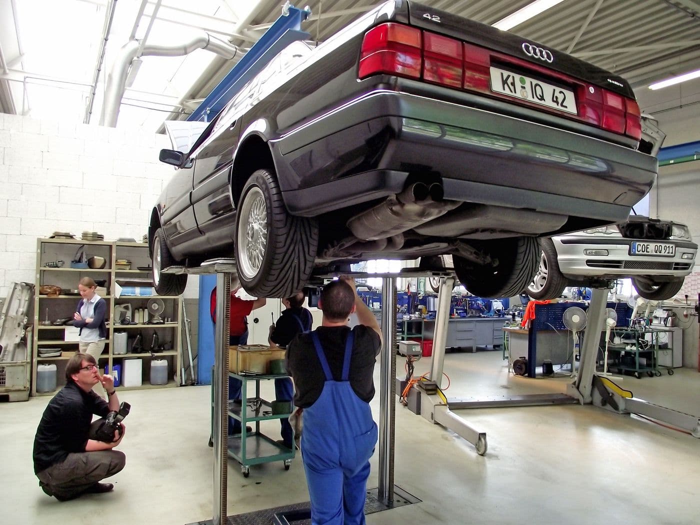 Blick unter den Audi v8 in der Werkstatt auf einer Hebebühne. Zwei Männer begutachten den Wagen von unten.