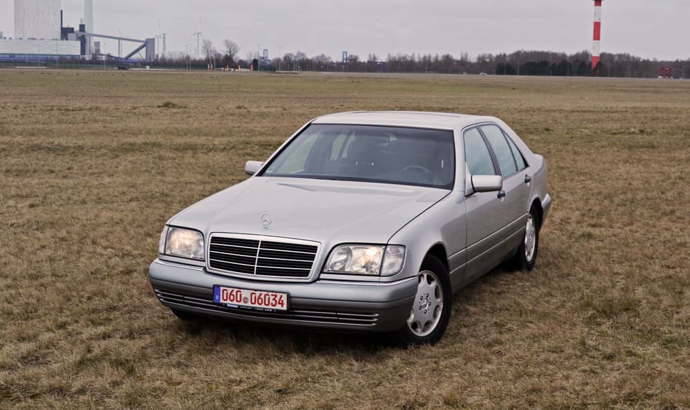 Frontansicht eines grauen Mercedes Benz w140 s320 parkend auf einer grünen Wiese mit Wald im Hintergrund