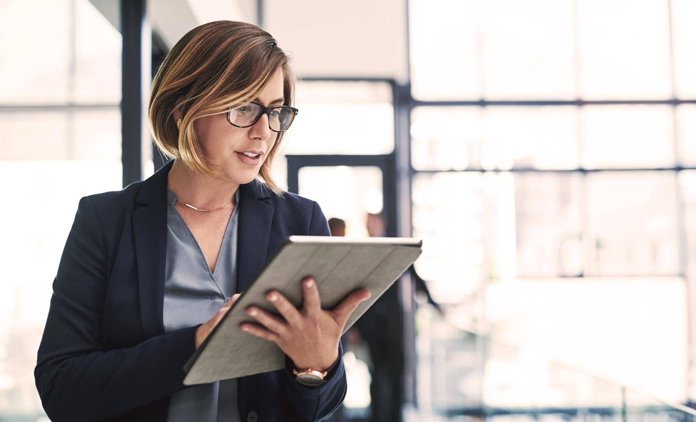 Eine Frau trägt einen dunkelblauen Blazer und eine Brille. Sie schaut auf ein Tablet.