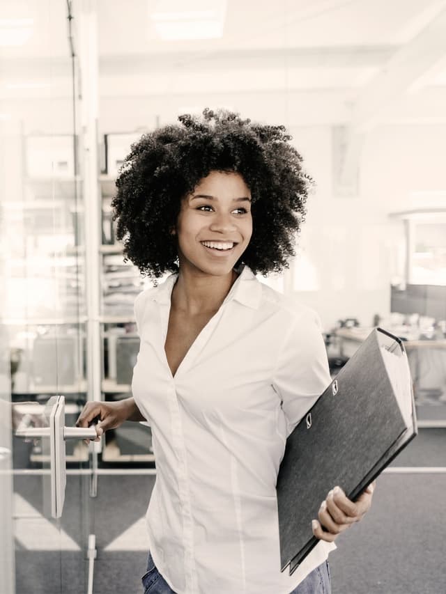 Eine Frau mit lockigem Haar lächelt und hält einen Ordner. Im Hintergrund ist ein Büro zu erkennen.