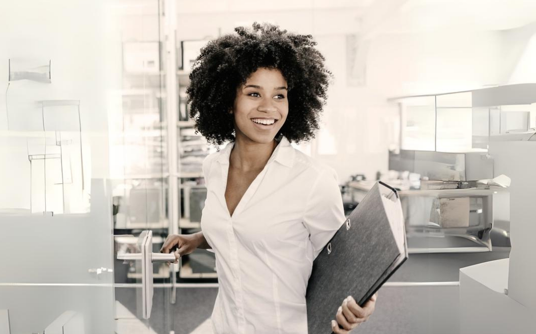 Junge Business-Frau mit Afro und weißem Hemd hält einen Artenordner und geht durch das Büro.