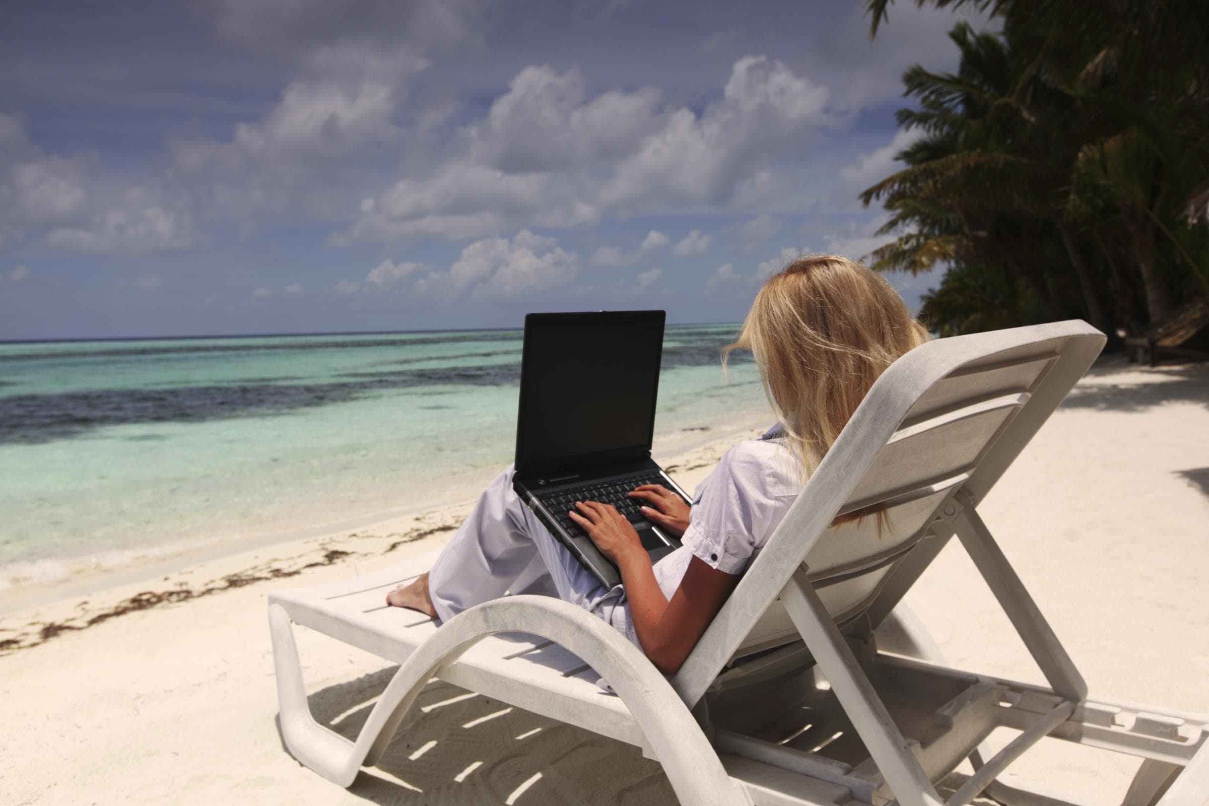 Frau sitzt auf einer Liege am Strand und arbeitet an einem Laptop, im Hintergrund das türkisfarbene Meer und Palmen