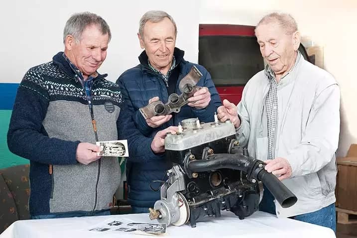 Drei ältere Männer stehen um einen Motor auf einem Tisch, begutachten Motorteile und halten alte Fotos in der Hand