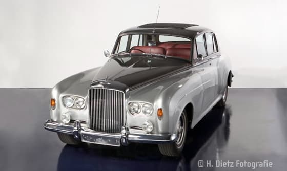 Ein klassischer Bentley in Silber und mit weinroten Sitzen wird in einem Studio präsentiert, umgeben von stilvollem Licht und eleganter Atmosphäre.