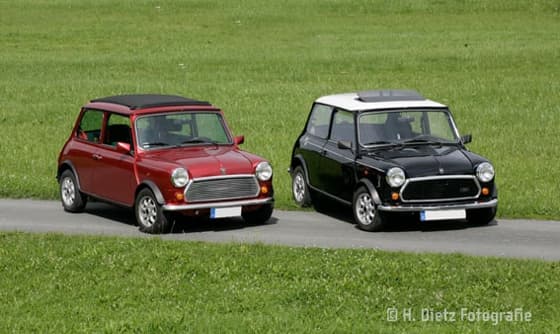 Zwei Oldtimer-Minis, ein roter und ein schwarzer, parken nebeneinander auf einem Weg.