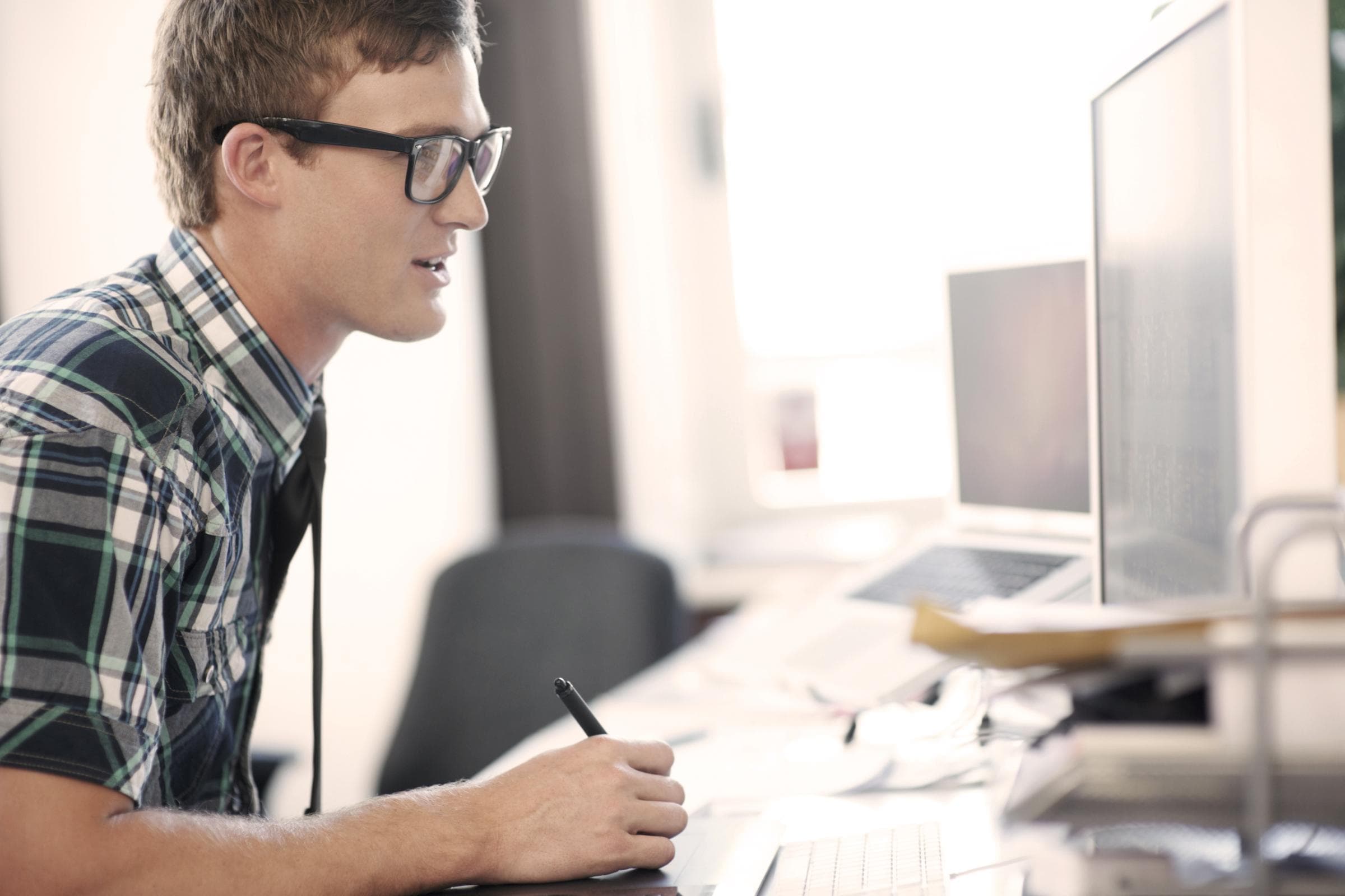 Ein junger Mann mit Brille sitzt an einem Schreibtisch vor einem Computerbildschirm. Er trägt ein kariertes Hemd und hält einen Eingabestift in der Hand. Im Hintergrund sind weitere technische Geräte und Arbeitsutensilien zu sehen.