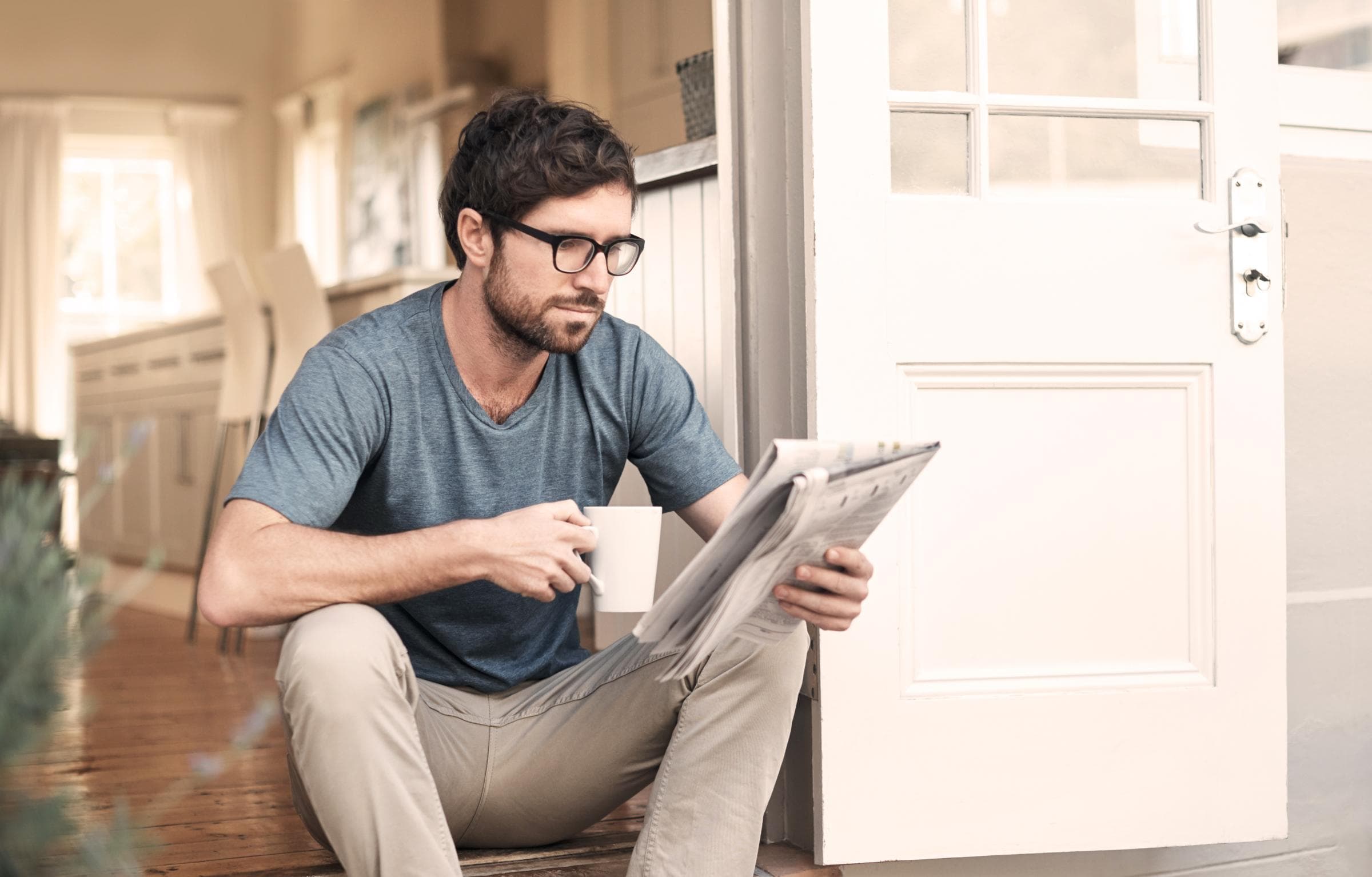 Ein Mann sitzt auf den Stufen seines Hauses, liest eine Zeitung und hält dabei eine Tasse in der Hand.