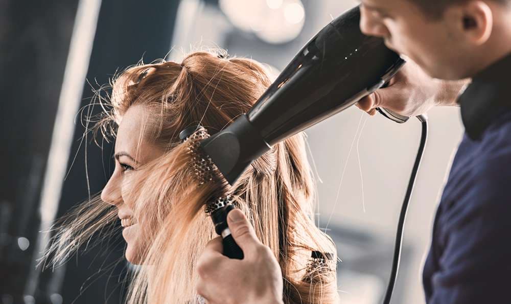 Friseur trocknet das Haar einer lächelnden Frau mit einem Föhn, während er eine Rundbürste benutzt