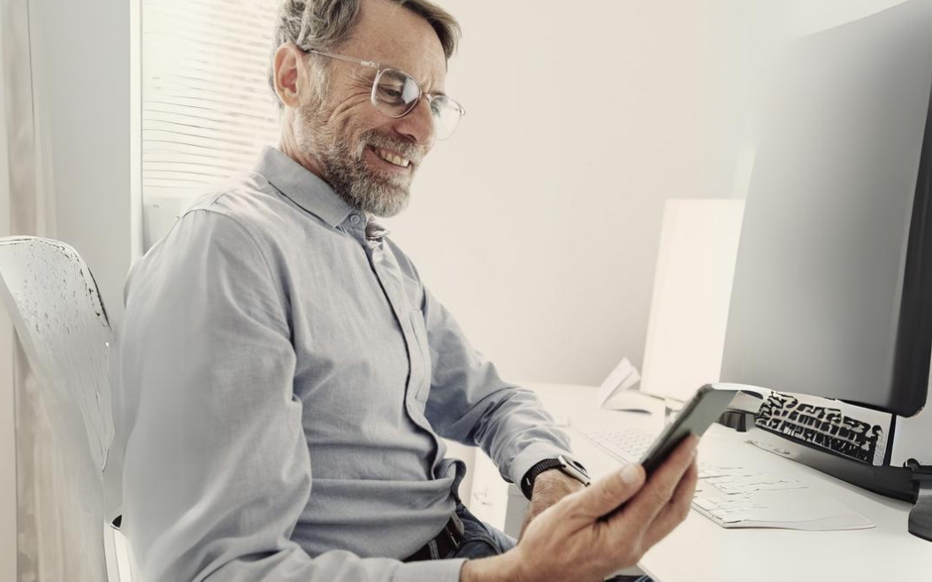 Business-Mann in hellblauem Hemd sitzt am Schreibtisch und hält sein Handy in der Hand.