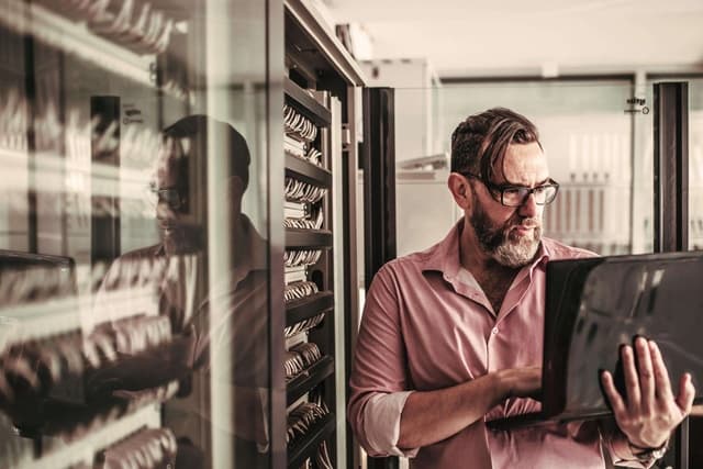 Mann mit Brille und Bart arbeitet an einem Laptop in einem Serverraum. Im Hintergrund sind mehrere Server-Racks zu sehen. Das Bild spiegelt sich in der Glaswand neben ihm.