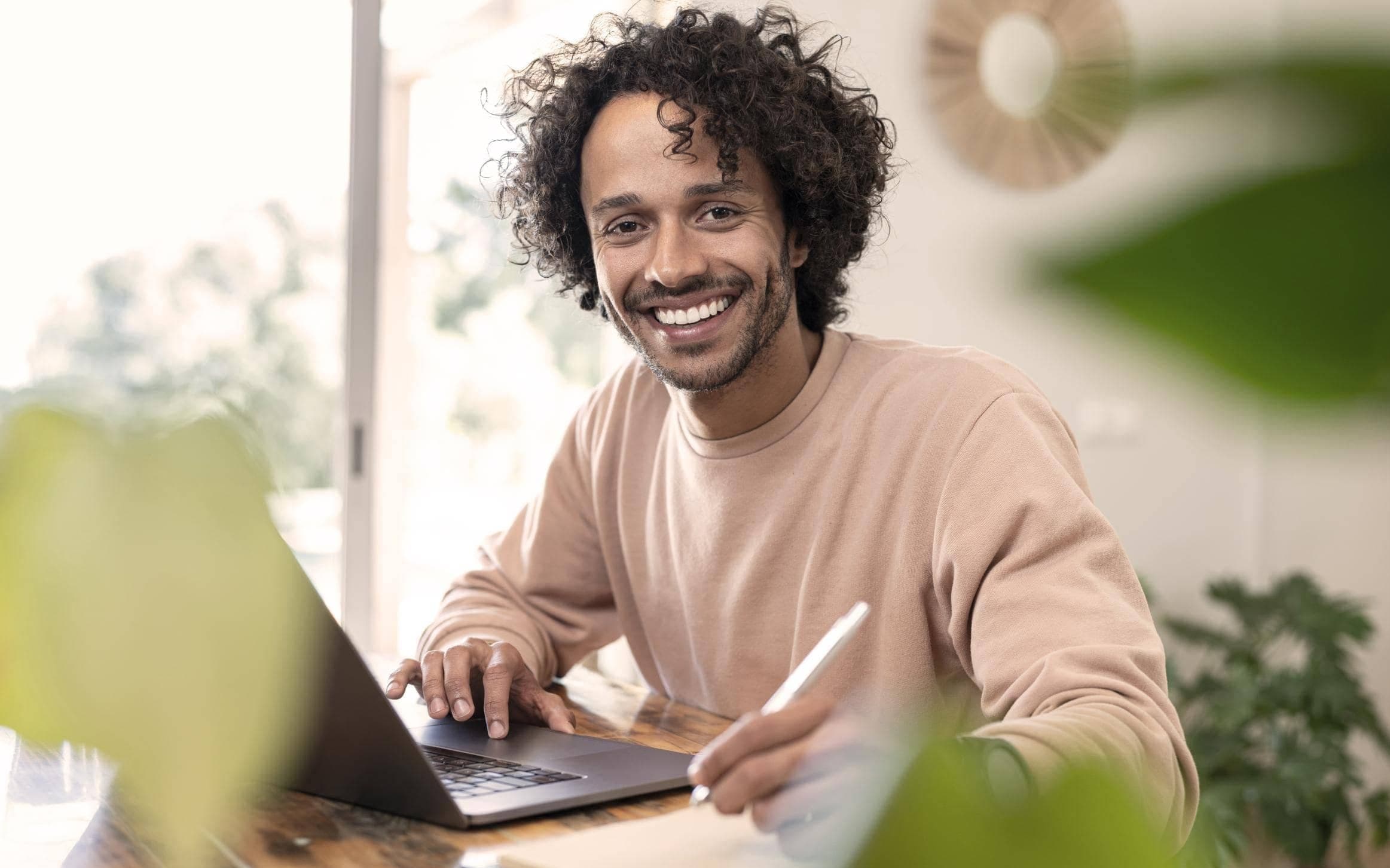 Junger Journalist mit lockigen brauen Haaren und lachsfarbenem Oberteil sitzt an einem Schreibtisch mit seinem Laptop und lächelt in die Kamera.