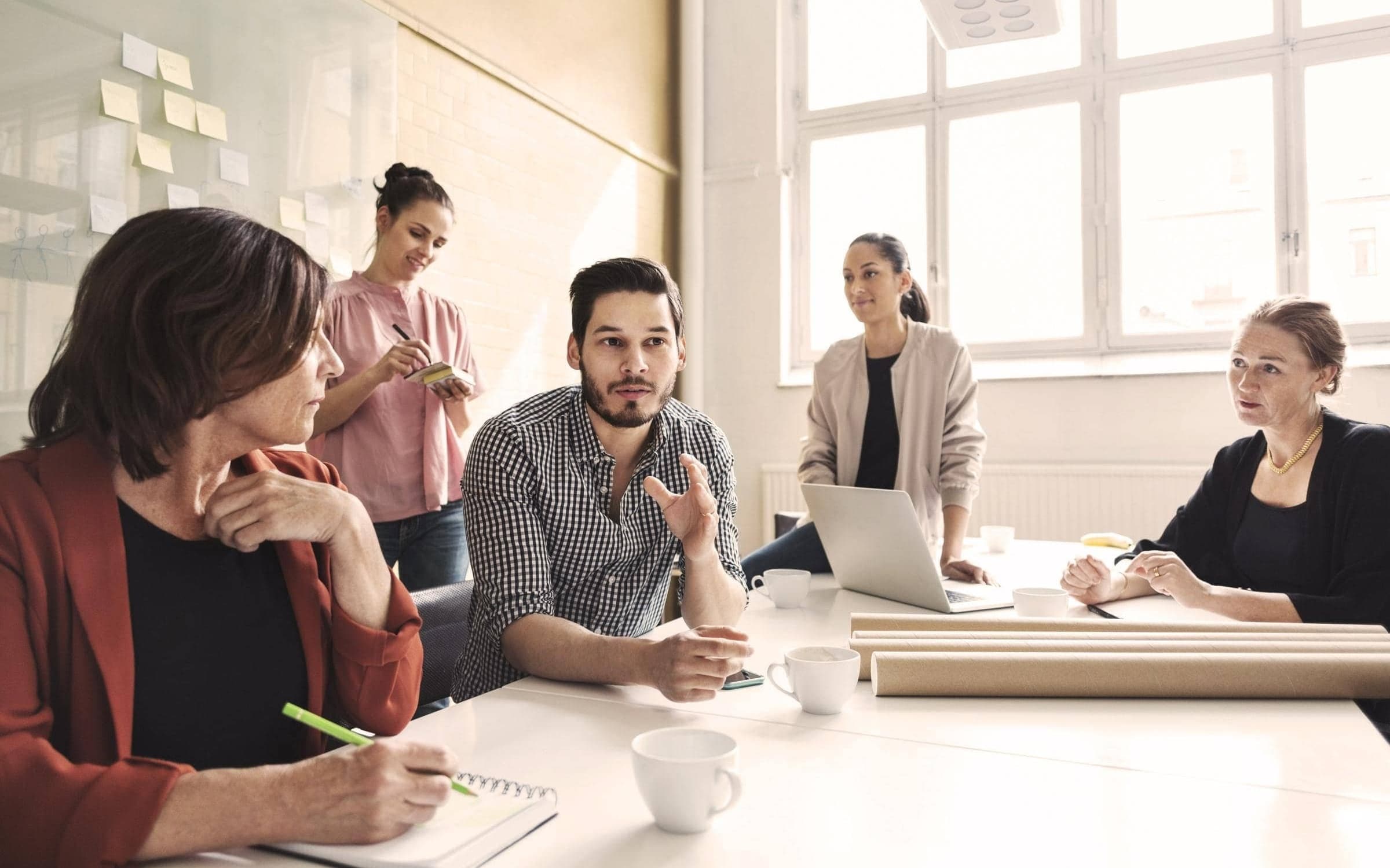 Junges fünfköpfiges Agenturteam sitzt in einem Meetingraum um einem Meetingtisch und diskutiert.