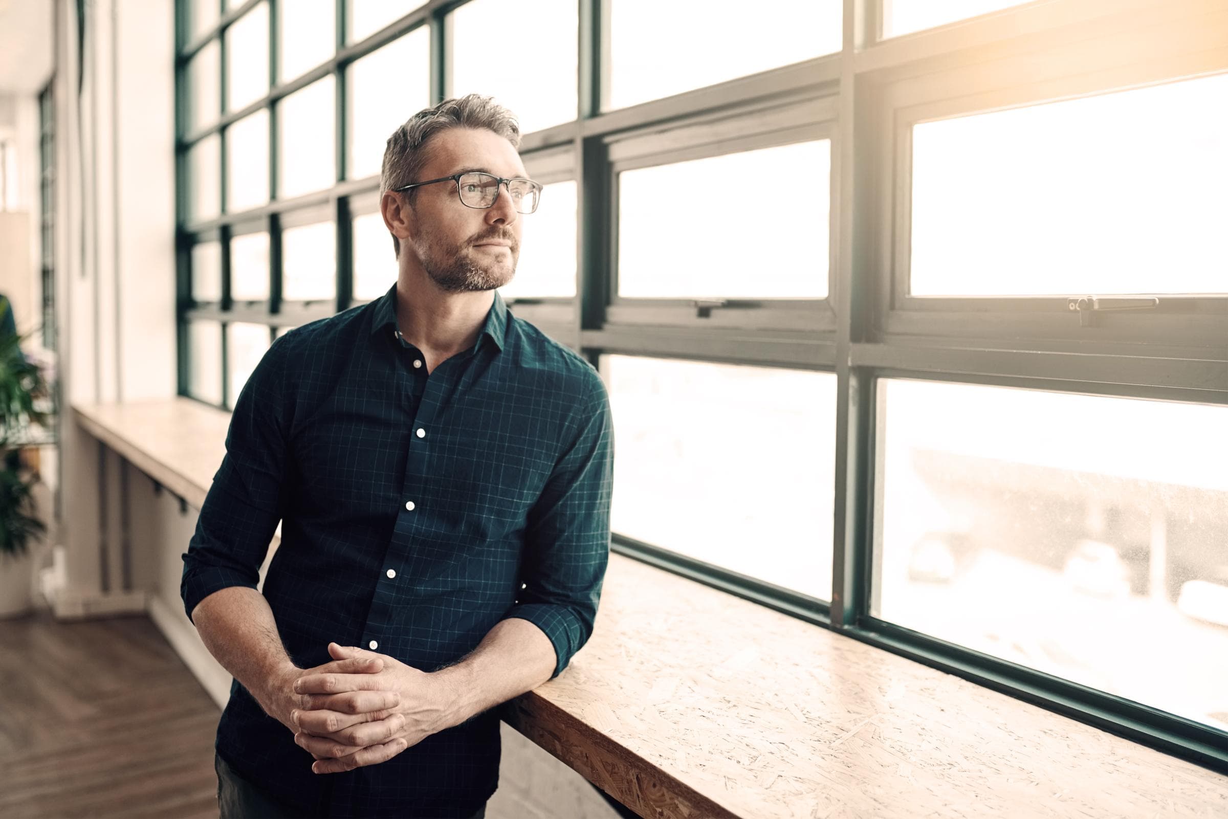 Ein Mann mit Brille und dunkelgrünem Hemd steht in einem modernen Büro und blickt nachdenklich aus einem grossen Fenster mit viel Tageslicht.