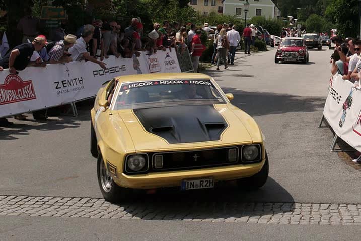 Gelber Ford Mustang fährt an einer Menschenmenge und Werbebannern vorbei während einer Oldtimer-Rallye