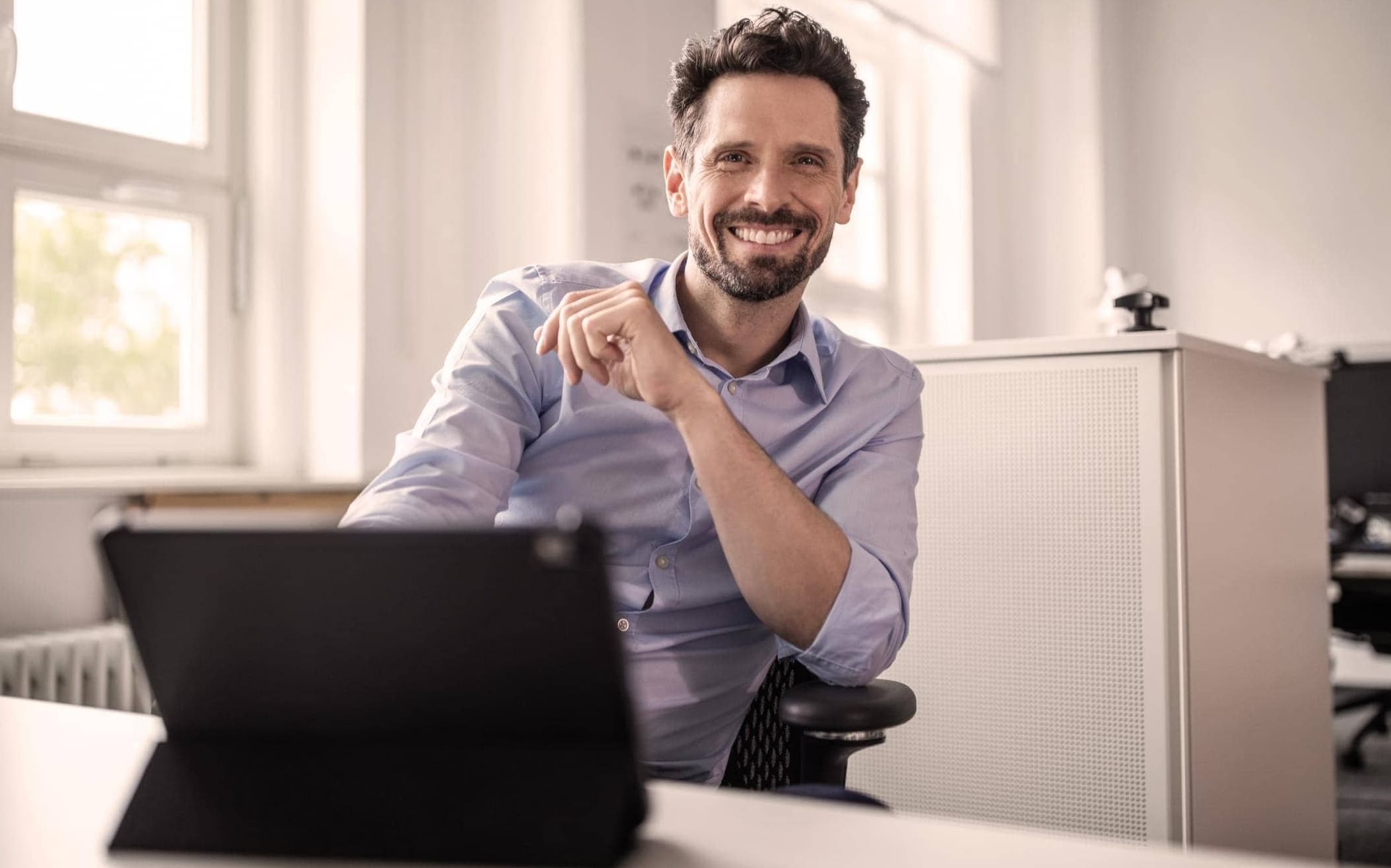 Unternehmensberater mit blauem Hemd und braunen Haaren sitzt vor seinem Tablet am Schreibtisch und lächelt in die Kamera.