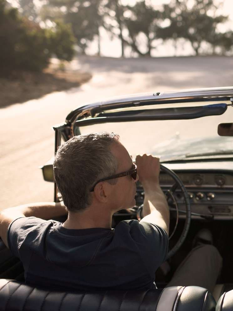 Mann sitzt in einem Oldtimer-Cabrio und fährt auf einer Straße. Er trägt eine Sonnenbrille, ein T-Shirt und hält das Lenkrad