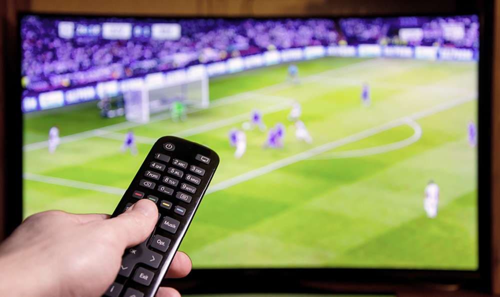Fußballspiel läuft in Fernseher und wird live übertragen. Hand hält eine Fernbedienung vor den TV.